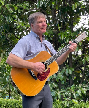 Gavin, standing before a leafy background, playing guitar, looking serious.