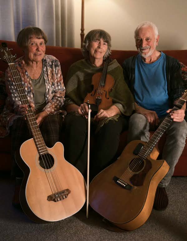 Jenny, Jude, & Tony, on a couch, Tony & Jenny are holding guitars, Jude her violin & bow